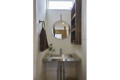 丸鏡がオシャレな手洗い場。ステンレスの洗面台と木製棚、漆喰の壁など、シンプルながら素材感が引き立つ空間です。
